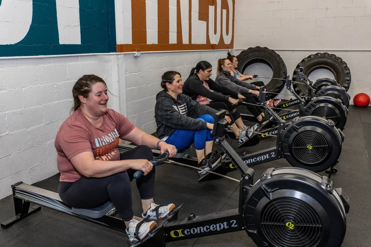 Members laughing together on rowing machines in front of the painted studio wall