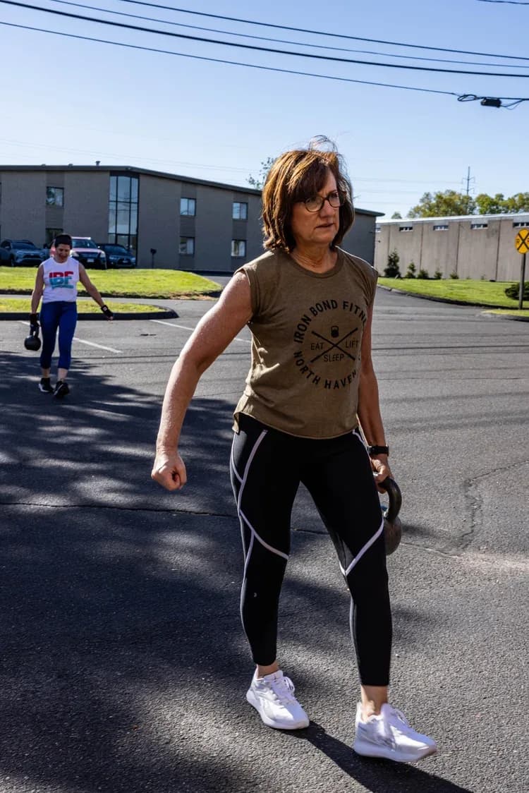 A member in a studio tee carrying two kettlebells outdoors